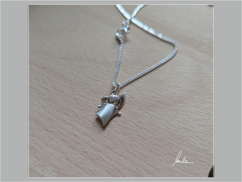 Peut inclure: Un collier avec pendentif argent&eacute; repr&eacute;sentant un ange stylis&eacute;. Le pendentif est suspendu &agrave; une cha&icirc;ne d&eacute;licate, pos&eacute; sur une surface en bois brun clair. L'ange a une robe fluide et des ailes d&eacute;ploy&eacute;es.
