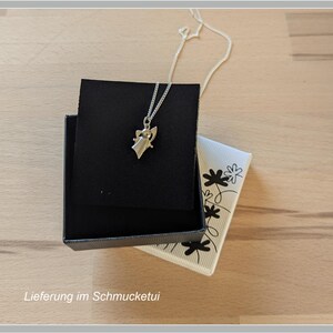 Peut inclure: Un collier pendentif argent&eacute; avec un petit ange est pos&eacute; sur un &eacute;crin noir. La bo&icirc;te est ouverte, r&eacute;v&eacute;lant une carte blanche &agrave; motifs floraux. Le texte "Lieferung im Schmucketui" est visible en bas.