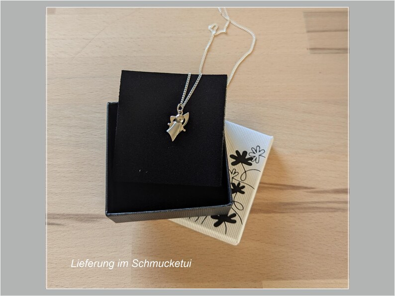 Peut inclure: Un collier pendentif argent&eacute; avec un petit ange est pos&eacute; sur un &eacute;crin noir. La bo&icirc;te est ouverte, r&eacute;v&eacute;lant une carte blanche &agrave; motifs floraux. Le texte "Lieferung im Schmucketui" est visible en bas.