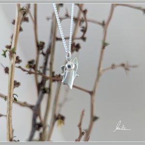 Peut inclure: Un collier pendentif argent&eacute; avec un charm en forme d'ange stylis&eacute;. L'ange a un design simple avec des ailes d&eacute;ploy&eacute;es et une petite t&ecirc;te. Le collier est suspendu devant un fond de branches brunes.