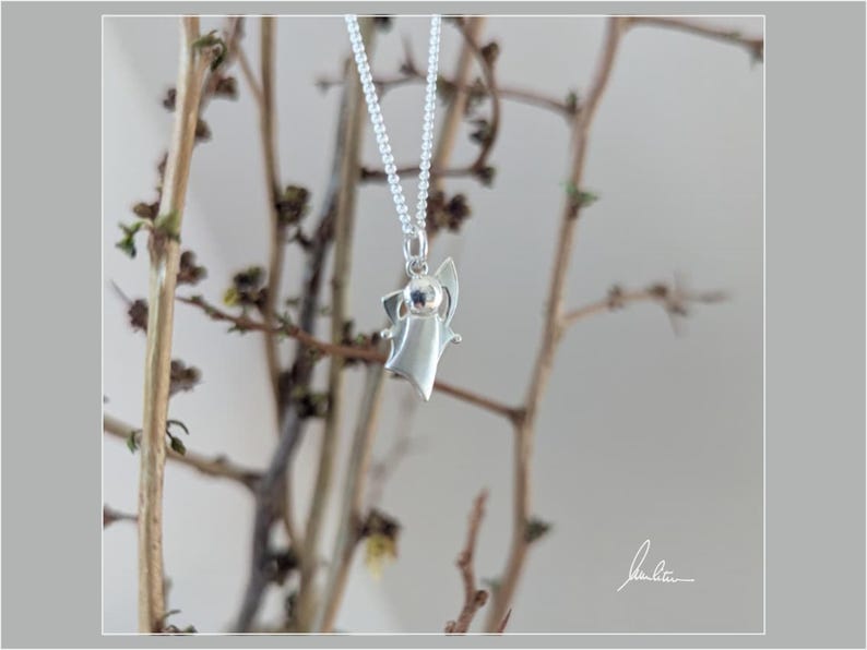 Peut inclure: Un collier pendentif argent&eacute; avec un charm en forme d'ange stylis&eacute;. L'ange a un design simple avec des ailes d&eacute;ploy&eacute;es et une petite t&ecirc;te. Le collier est suspendu devant un fond de branches brunes.