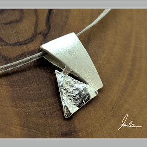 Peut inclure: Un collier pendentif argenté au design géométrique. Le pendentif présente deux formes triangulaires reliées, l'une lisse et l'autre texturée, suspendues à une fine chaîne argentée. Le collier est présenté sur une surface en bois.