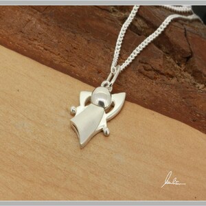 Peut inclure: Un collier avec pendentif ange en argent. Le pendentif repr&eacute;sente un ange avec une t&ecirc;te arrondie, un corps fluide et des formes en forme d'ailes. La cha&icirc;ne du collier est une fine cha&icirc;ne en argent. Le pendentif et la cha&icirc;ne sont pr&eacute;sent&eacute;s sur une surface en bois.