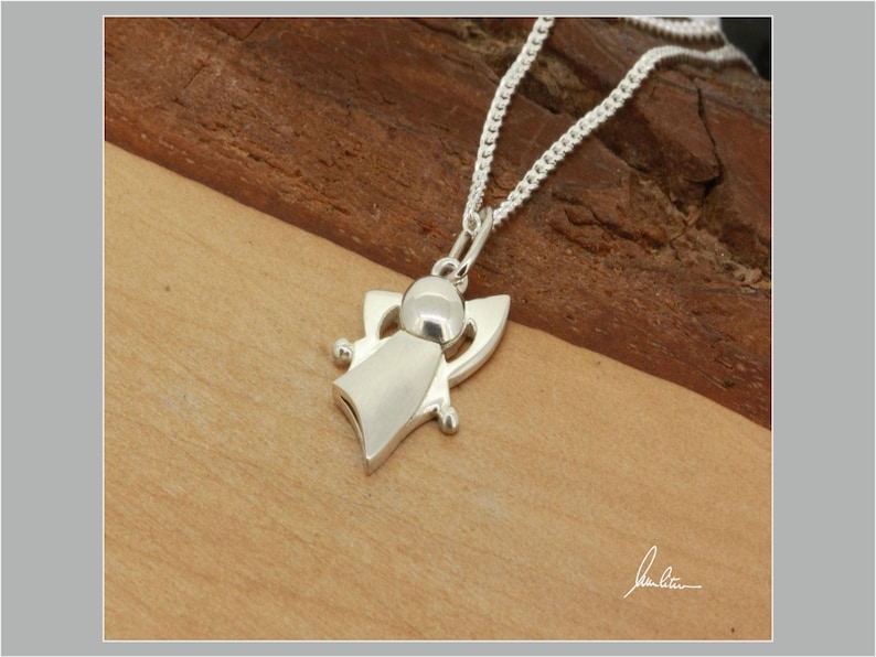 Peut inclure: Un collier avec pendentif ange en argent. Le pendentif repr&eacute;sente un ange avec une t&ecirc;te arrondie, un corps fluide et des formes en forme d'ailes. La cha&icirc;ne du collier est une fine cha&icirc;ne en argent. Le pendentif et la cha&icirc;ne sont pr&eacute;sent&eacute;s sur une surface en bois.