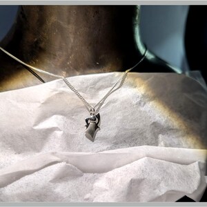 Peut inclure: Un collier pendentif en argent avec un design g&eacute;om&eacute;trique. Le pendentif est suspendu &agrave; une cha&icirc;ne fine. Le collier est pr&eacute;sent&eacute; sur une surface blanche, avec un fond sombre.