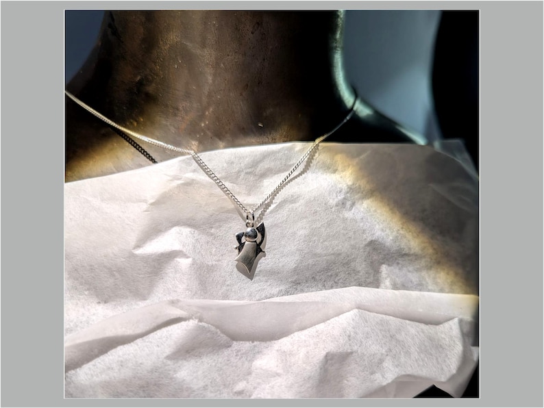Peut inclure: Un collier pendentif en argent avec un design g&eacute;om&eacute;trique. Le pendentif est suspendu &agrave; une cha&icirc;ne fine. Le collier est pr&eacute;sent&eacute; sur une surface blanche, avec un fond sombre.