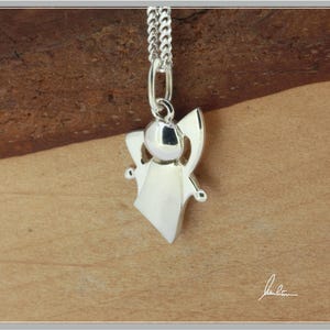 Peut inclure: Pendentif ange argent&eacute; suspendu &agrave; une cha&icirc;ne argent&eacute;e. Le pendentif pr&eacute;sente une t&ecirc;te ronde, des ailes d&eacute;ploy&eacute;es et une robe fluide. Le collier est pr&eacute;sent&eacute; sur un fond en bois chaleureux.