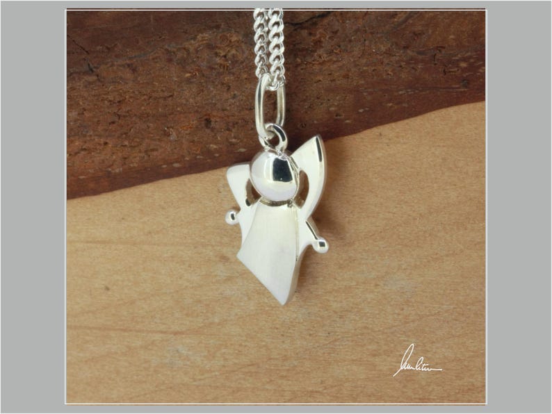 Peut inclure: Pendentif ange argent&eacute; suspendu &agrave; une cha&icirc;ne argent&eacute;e. Le pendentif pr&eacute;sente une t&ecirc;te ronde, des ailes d&eacute;ploy&eacute;es et une robe fluide. Le collier est pr&eacute;sent&eacute; sur un fond en bois chaleureux.