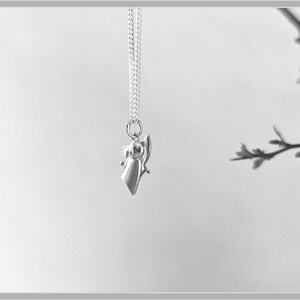 Peut inclure: Un collier pendentif argent&eacute; avec un petit ange stylis&eacute;. L'ange a une robe fluide et des ailes d&eacute;ploy&eacute;es, suspendu &agrave; une cha&icirc;ne d&eacute;licate. Le fond est d'une tonalit&eacute; neutre douce, mettant en valeur le bijou.