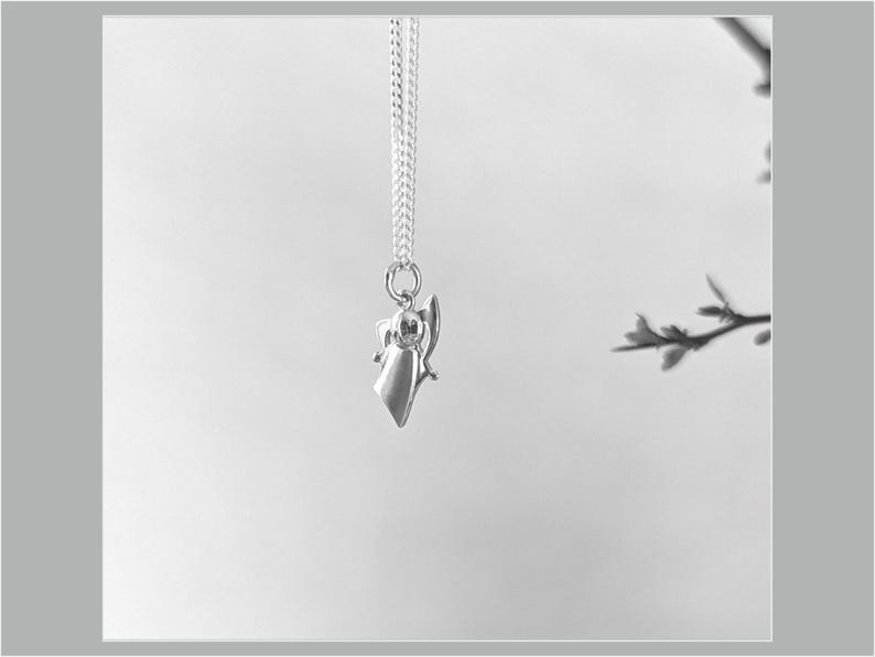 Peut inclure: Un collier pendentif argent&eacute; avec un petit ange stylis&eacute;. L'ange a une robe fluide et des ailes d&eacute;ploy&eacute;es, suspendu &agrave; une cha&icirc;ne d&eacute;licate. Le fond est d'une tonalit&eacute; neutre douce, mettant en valeur le bijou.