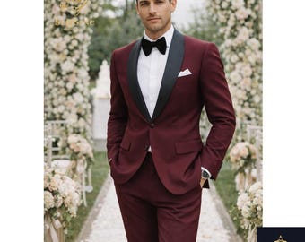 May include: A burgundy suit with a black lapel and a black bow tie. The suit has a white dress shirt and a white pocket square. The man is standing in front of a floral archway.