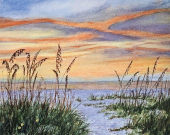 May include: Watercolor painting of a beach scene at sunset. The sky transitions from blue to orange and yellow. Tall grasses and green plants frame a sandy path leading to the ocean. The text "Barbara Rosenzweig Watercolors" is visible.
