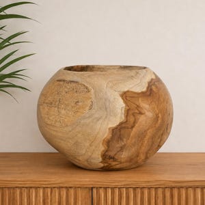 Puede incluir: Un cuenco de madera redondo y hueco con un patrón de vetas naturales en tonos marrón claro y oscuro. El cuenco tiene una parte superior ancha y abierta, y una superficie exterior lisa y curva que muestra la textura orgánica de la madera.