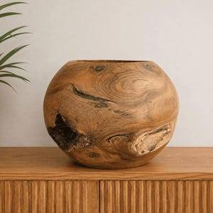 Peut inclure: Un bol sphérique en bois naturel, présentant un motif de grain prononcé avec des nœuds visibles et des inclusions organiques sombres et texturées. La surface affiche des nuances variées de brun clair et moyen, révélant la texture brute du bois.