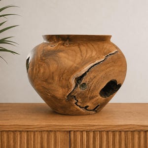 Peut inclure: Un vase en bois arrondi sculpté dans une seule pièce de bois brun clair, doté d'une large ouverture sur le dessus et d'une fissure naturelle sombre et irrégulière ou d'une section creuse sur sa face avant, révélant le grain du bois.