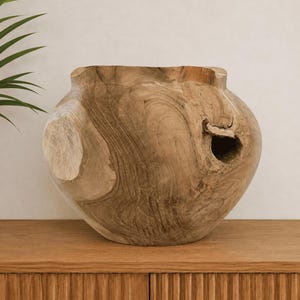 Peut inclure: Un grand récipient en bois arrondi, sculpté dans une seule pièce de bois brun clair. La surface présente des motifs de grain de bois naturels, des nœuds et une ouverture organique distincte sur le côté, révélant la texture brute du matériau.