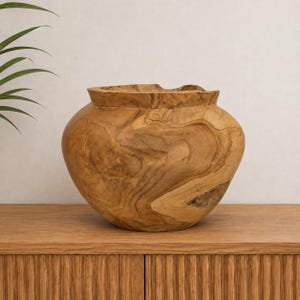 Puede incluir: Un cuenco redondeado de madera de teca natural, con una abertura ancha y un patrón de veta distintivo con tonos arremolinados en marrón claro y oscuro. El cuenco tiene una superficie lisa y pulida y un borde grueso con textura orgánica.