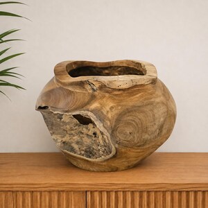 Peut inclure: Un grand récipient en bois arrondi sculpté dans une seule pièce de bois naturel. L'extérieur présente un grain brun clair riche avec des nœuds visibles, des textures organiques et un centre évidé avec un bord large, irrégulier et épais.