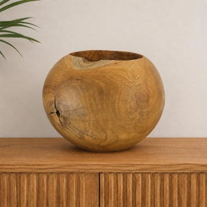 Puede incluir: Un cuenco esférico de madera fabricado en madera clara natural, con un prominente patrón de veta orgánica, un exterior redondeado y liso, una amplia abertura circular en la parte superior y un nudo natural visible en la superficie lateral.