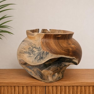 Puede incluir: Un cuenco grande de madera de teca ahuecada con una forma natural y orgánica. El exterior presenta una mezcla de tonos de madera marrón cálido y secciones de color gris carbón oscuro, mostrando la textura cruda e irregular de la raíz.