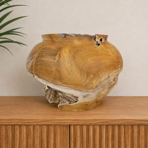 Può includere: Una ciotola in legno di teak naturale con un corpo largo e arrotondato e un'apertura stretta. L'esterno presenta una venatura del legno pronunciata in tonalità marrone dorato e beige, con bordi grezzi e testurizzati e nodi organici sul bordo.