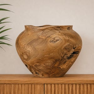 Puede incluir: Un jarrón grande y redondeado tallado en madera natural de color marrón claro, con una abertura ancha en la parte superior y una base estrecha. La superficie muestra vetas de madera prominentes, nudos en espiral y pequeñas grietas naturales.