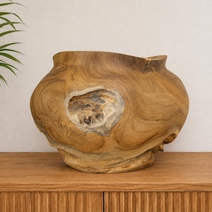 Peut inclure: Un grand bol en bois arrondi, sculpté dans une seule pièce de bois de teck brun clair. Le bol présente un nœud texturé proéminent au centre avec des motifs de grain tourbillonnants et un large sommet ouvert au bord irrégulier et organique.