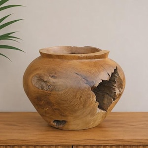 Op de afbeelding: Een afgeronde houten vaas gesneden uit één stuk lichtbruin hout, met een brede opening aan de bovenkant, een glad gepolijst buitenoppervlak en een grote, grillige, natuurlijke opening aan de zijkant die de ruwe houtnerf blootlegt.
