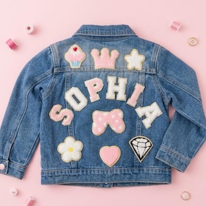 Puede incluir: Chaqueta vaquera azul con el nombre "SOPHIA" aplicado en la espalda en letras rosas y blancas. La chaqueta presenta varios parches, incluyendo una magdalena, una corona, una estrella, un lazo, una flor, un corazón y un diamante.