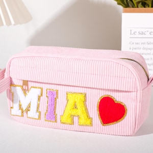May include: A pink corduroy cosmetic bag with the name "MIA" in colourful, embroidered letters and a red heart. The bag has a zip closure and small handles. The bag is rectangular and has a vertical ribbed texture.