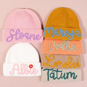 May include: A stack of six knitted beanies in various colors, including pink, white, and mustard yellow. Each beanie features a name embroidered in a contrasting color. The names include "Sloane", "Allie", "Mareya", "Vedha", "Lorelei", and "Tatum".