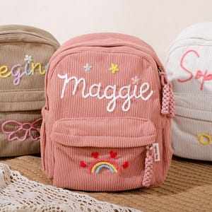 Peut inclure: Trois sacs à dos en velours côtelé beige, rose et blanc, chacun brodé d'un nom. Le sac à dos rose présente un motif arc-en-ciel et cœur. Les sacs ont des poches avant et des pompons décoratifs. Un accessoire mignon et personnalisé.