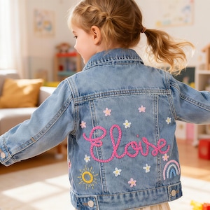 May include: Light blue denim jacket with embroidered pink flowers, a sun, a rainbow, and the name "Eloise" in pink thread. The jacket is a classic style, perfect for children's wear.