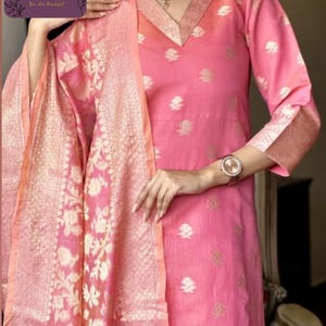 May include: Pink Indian kurta suit with a matching dupatta. The kurta features a V-neck and floral pattern. The dupatta has a gold floral design. The outfit is made of a lightweight fabric.