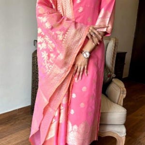 May include: A pink salwar kameez suit with gold floral embroidery. The outfit includes a long-sleeved top, matching pants, and a dupatta. The dupatta and sleeves have gold patterned borders. The outfit is worn with a watch.