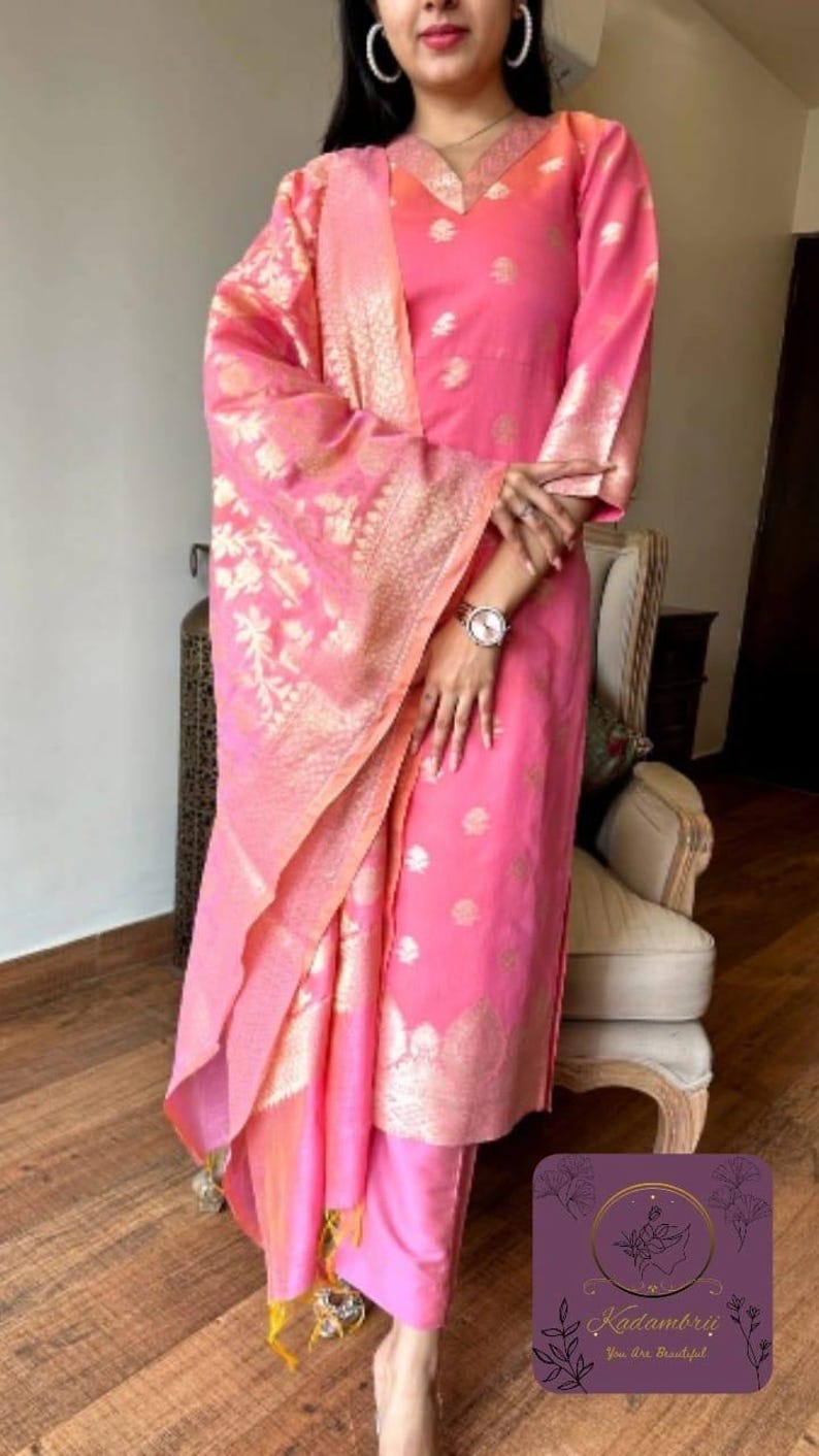 May include: A pink salwar kameez suit with gold floral embroidery. The outfit includes a long-sleeved top, matching pants, and a dupatta. The dupatta and sleeves have gold patterned borders. The outfit is worn with a watch.