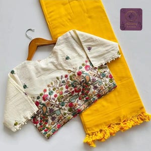 May include: Cream-colored embroidered blouse with long sleeves, featuring a vibrant floral and bird design in pink, green, and blue. Paired with a bright yellow fabric with a fringe trim. The blouse is hanging on a wooden hanger.