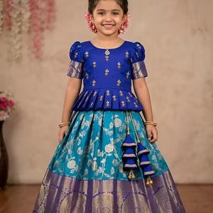 May include: A young girl wearing a vibrant blue and turquoise traditional Indian dress. The top is royal blue with gold embroidery and short sleeves. The skirt is turquoise with a floral pattern and a purple border. Gold tassels hang from the waist.