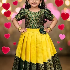 May include: A young girl wearing a traditional Indian dress. The dress features a green floral patterned top with short sleeves and a yellow pleated skirt with green and gold patterned trim. Gold bangles adorn her wrists.