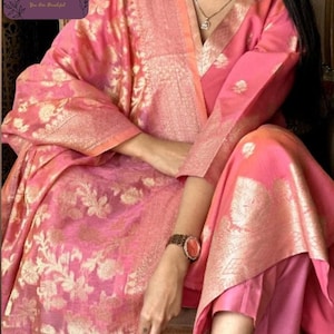 May include: A pink and gold floral patterned salwar kameez ensemble. The outfit includes a long-sleeved tunic, matching pants, and a dupatta. The fabric has a shimmering effect, and the design features intricate floral patterns. The outfit is suitable for special occasions.