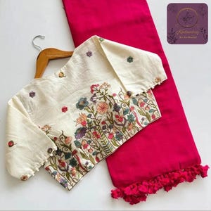 May include: An off-white blouse with long sleeves, adorned with colorful floral embroidery, is displayed on a wooden hanger. It is paired with a vibrant fuchsia-colored fabric, possibly a skirt or pants, with a tassel trim. The ensemble is set against a white background.