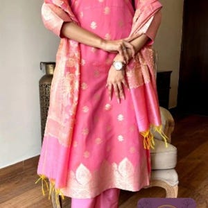 May include: A pink Indian salwar kameez suit with gold accents. The outfit includes a long tunic, matching pants, and a shawl. The tunic and shawl have gold floral patterns. The outfit is worn with embellished heels.