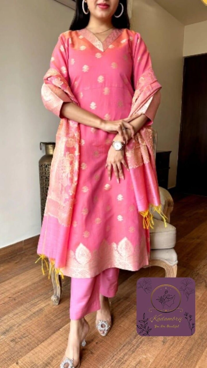 May include: A pink Indian salwar kameez suit with gold accents. The outfit includes a long tunic, matching pants, and a shawl. The tunic and shawl have gold floral patterns. The outfit is worn with embellished heels.