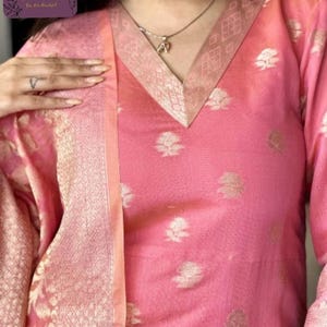 May include: A pink tunic-style top with a V-neck and floral pattern. The neckline and sleeves have a gold-colored trim. A matching scarf is draped over the shoulder. The top is part of a traditional outfit.