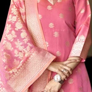 May include: Pink salwar kameez with gold floral embroidery and a matching dupatta. The outfit features a V-neck and three-quarter sleeves. The dupatta has a gold border. The outfit is adorned with small gold floral patterns.