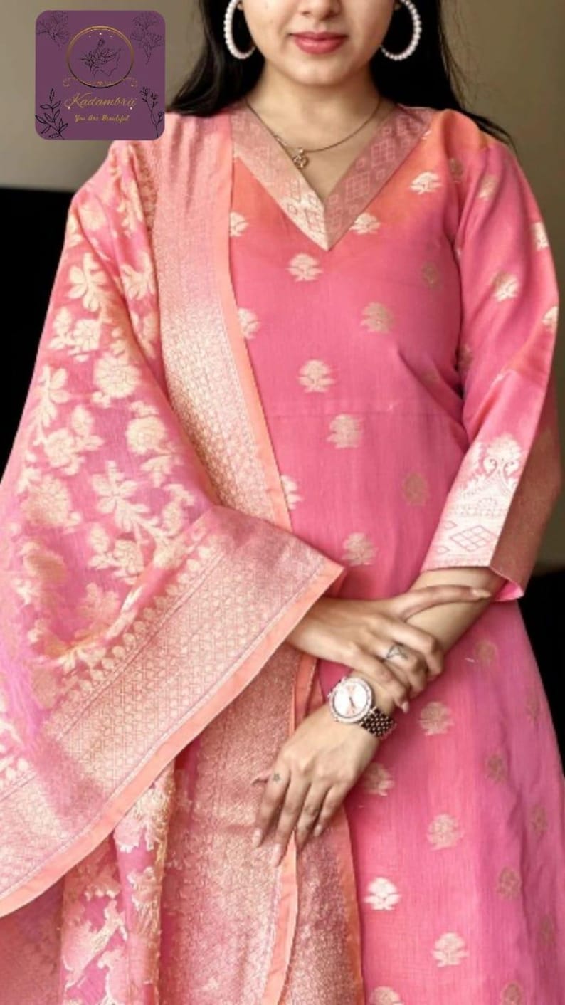 May include: Pink salwar kameez with gold floral embroidery and a matching dupatta. The outfit features a V-neck and three-quarter sleeves. The dupatta has a gold border. The outfit is adorned with small gold floral patterns.