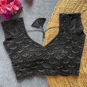 Puede incluir: Un top negro con cuentas y un escote en V profundo. El top sin mangas presenta un intrincado trabajo de cuentas en un patrón de vieira en el corpiño. Se ve una corbata negra en la espalda. El top se muestra sobre una superficie neutra.