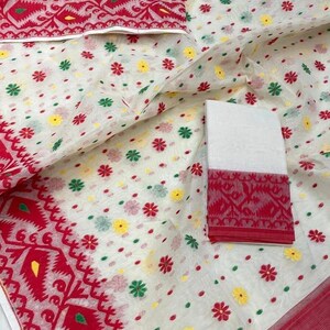 May include: White fabric with a red border and floral patterns. The fabric features small, colorful flower designs in red, yellow, and green. A folded section of the fabric displays a detailed red border with geometric patterns. The overall design is intricate and vibrant.