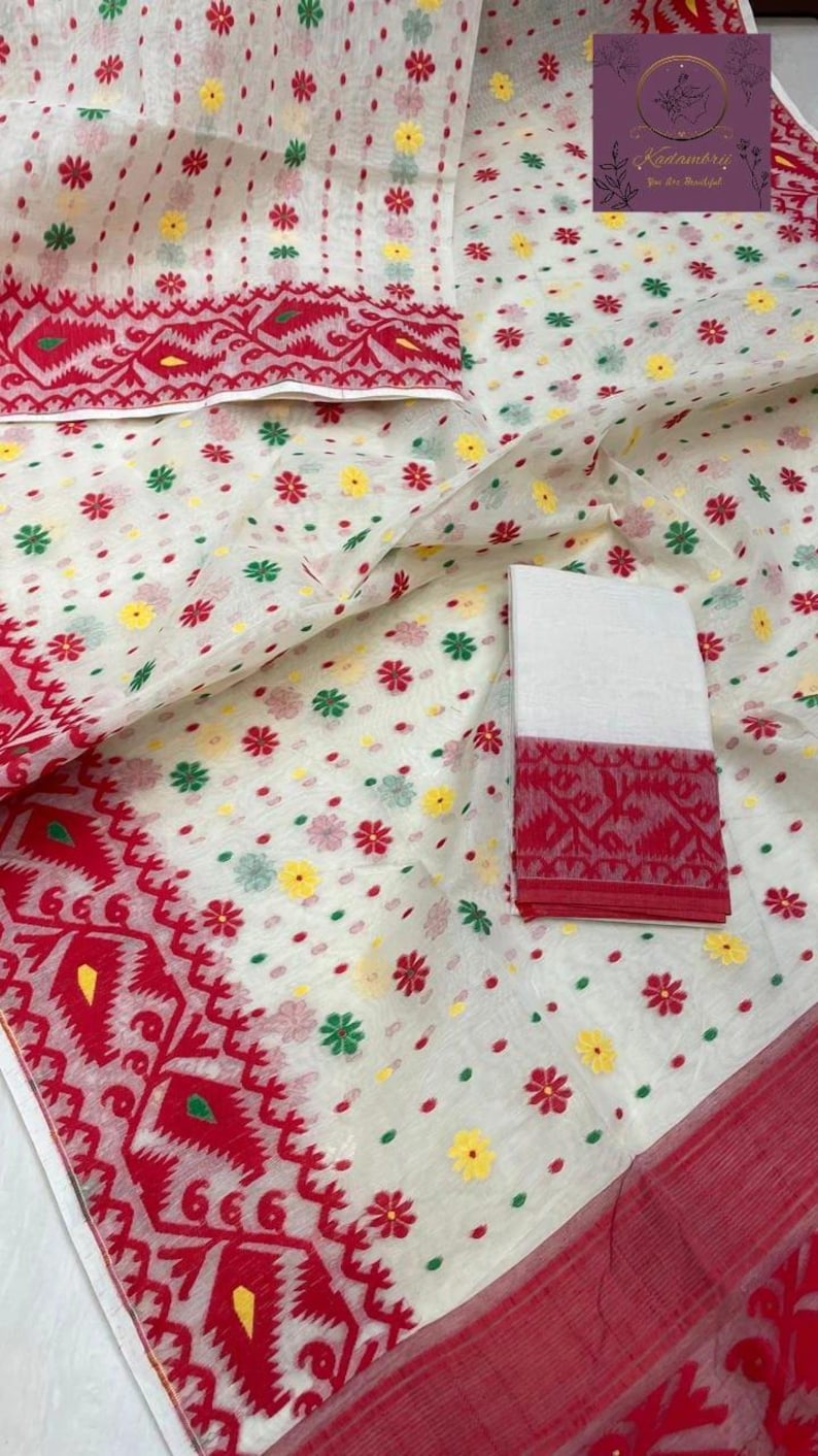May include: White fabric with a red border and floral patterns. The fabric features small, colorful flower designs in red, yellow, and green. A folded section of the fabric displays a detailed red border with geometric patterns. The overall design is intricate and vibrant.