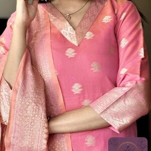 May include: Pink Indian kurta suit with gold floral patterns. The suit features a V-neckline and three-quarter sleeves. The dupatta and sleeve cuffs have a gold geometric design. The suit has the text "Kadambrii You are Beautiful" in the bottom right corner.